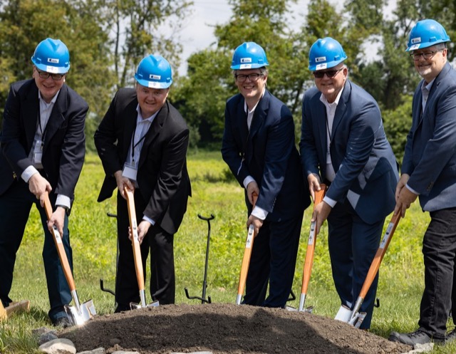 Five people in suits and blue hard hats turning soil with shovels at a groundbreaking ceremony